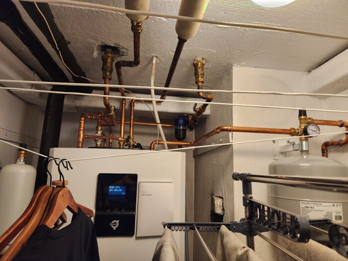 Tvättstuga med bergvärmepump, rörsystem och kopparledningar i taket. Klädhängare och torkställning under installationen.
