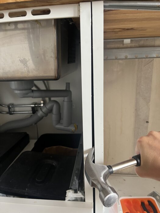 Hand håller hammare inuti köksskåp under diskbänk, visar rörsystem och återvinningsbehållare. Skåpstommen behöver demonteras.