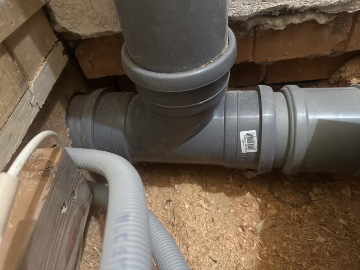Avloppsrör och flexibelt kabelrör installerade under ett hus, omgivet av trä och tegelväggar med sågspån på golvet.