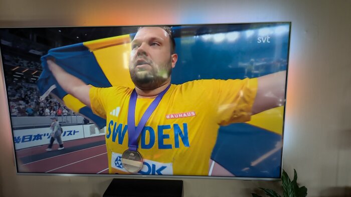 Svensk idrottare firar med en blågul flagga och medalj i gul tröja med texten "SWEDEN" på en arena.