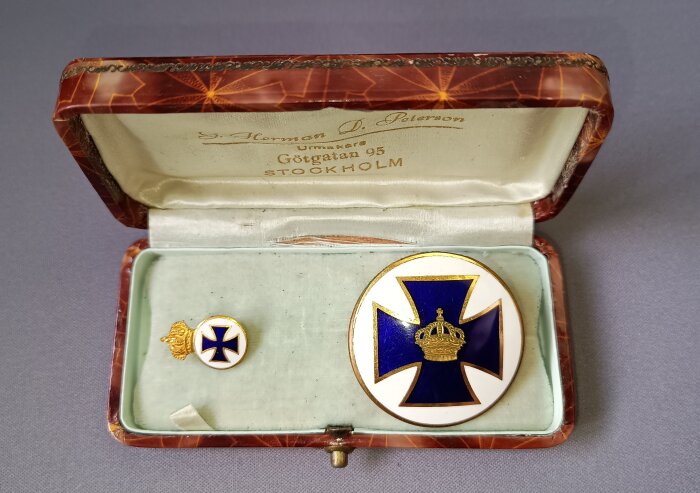 Brosch och nålmärke med kunglig krona och blått kors i etui från Götgatan, Stockholm.
