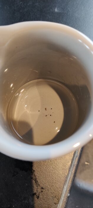 Beige mugg sedd ovanifrån med några små svarta och bruna fläckar inuti, placerad på en mörk yta.
