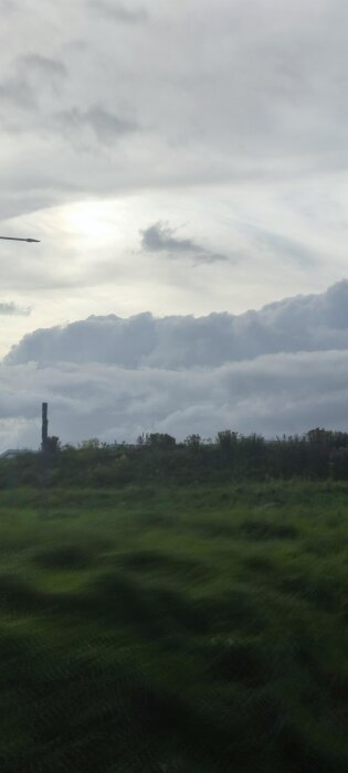Grön åker under molnig himmel med lågt stående sol, och en låg byggnad eller struktur i horisonten.