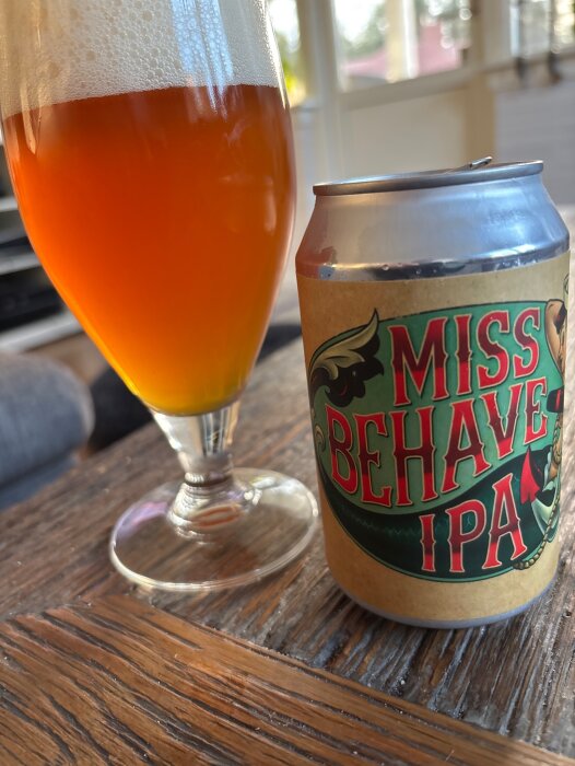 En ölburk med texten "Miss Behave IPA" bredvid ett glas med gyllene öl, stående på ett träbord.