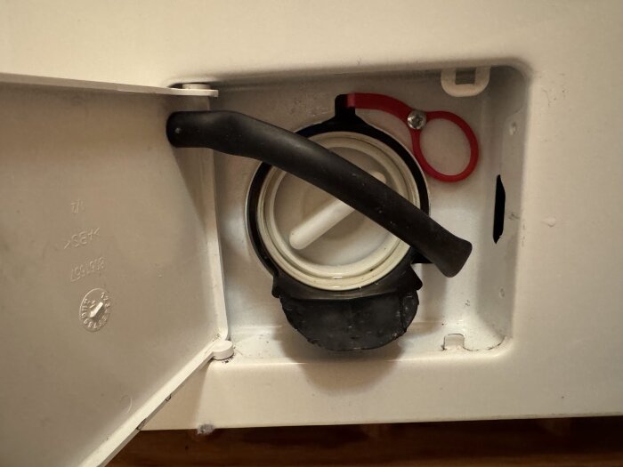 Bild på tvättmaskinens renslucka med en svart slang och röd nödöppningsring.