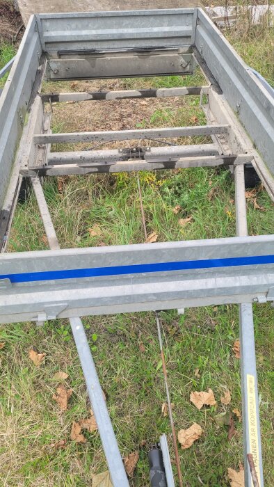 Ram av en släpvagn med avsaknad skiva, exponerade tvärgående balkar syns. Gräs och löv ligger under släpvagnen.