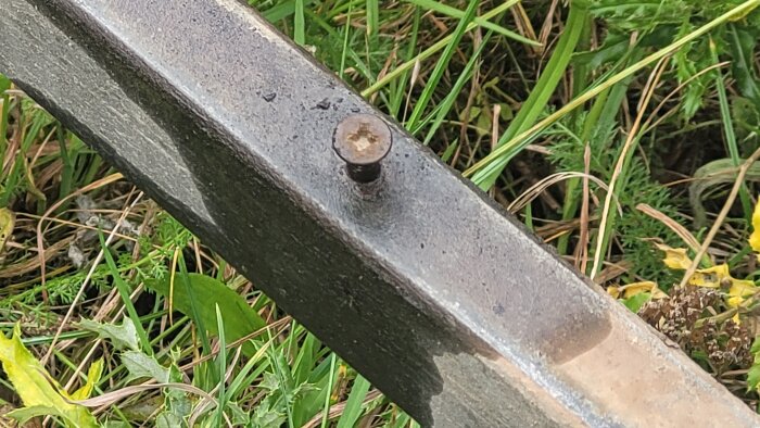 Närbild av en snedställd, rostig skruv i en metallbalk på gräsmark.