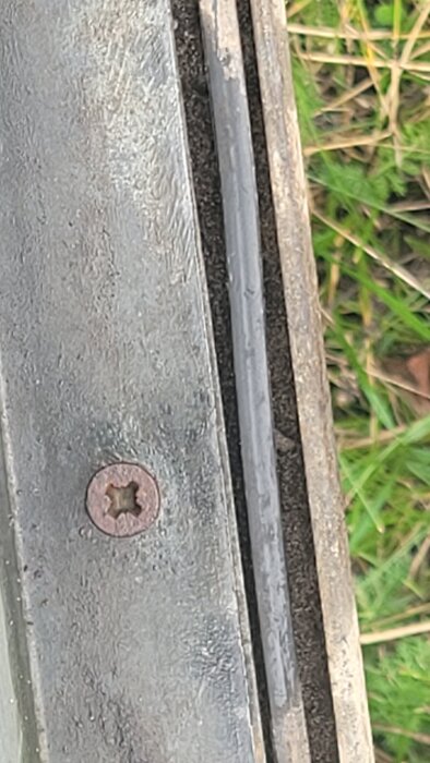 Närbild av metallskruv fastsatt i en plåt vid kanten av en släpvagnsram, med gräs i bakgrunden.