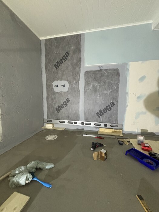 Vägg med uppmätt långpass, redo för kakling. Verktyg och material ligger spridda på golvet i ett rum under renovering.