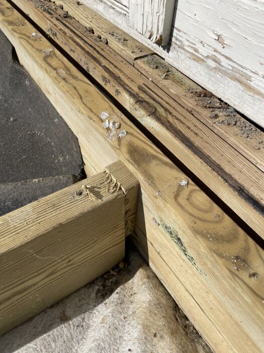 Två träreglar skarvas ihop intill ett slitet, målat träparti, omgivna av smuts och fukt, med några isflisor på ytan.