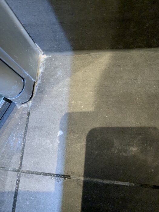 Fukt och vita fläckar vid golvtröskeln i badrum, potentiellt orsakade av byggfel eller fuktproblem, observerade på plattor och vägg.