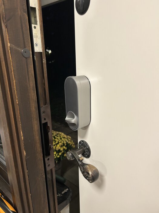 En vit dörr med ett installerat smartlås från Aqara och ett antikt dörrhandtag, delvis öppen mot en kvällsscen med växter.