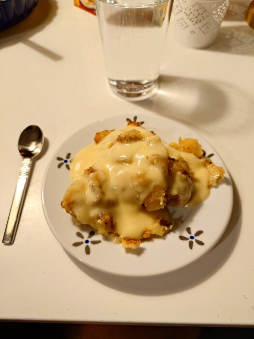 Äppelpaj med vaniljsås serverad på en blommig tallrik bredvid ett glas vatten och en sked på ett bord.