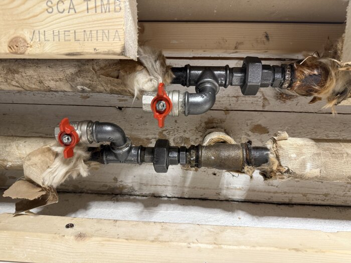 Avgrening med rör och ventiler kopplad till huvudledning för radiator i källare, med isolering och synliga träbjälkar i bakgrunden.
