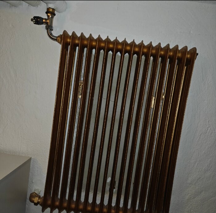 Gammal radiator mot vit vägg med anslutande värmerör synliga överst.