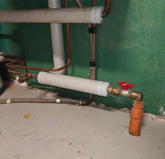 Rörsystem med flera koppar- och plastledningar, ventil med röd handtag, placerade mot en grön vägg och betonggolv.