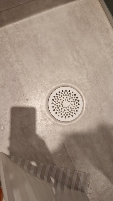 Närbild på badrumsgolv med ett runt vitt golvbrunnslock och tecken på blåsor under mattan i ett nyrenoverat badrum.