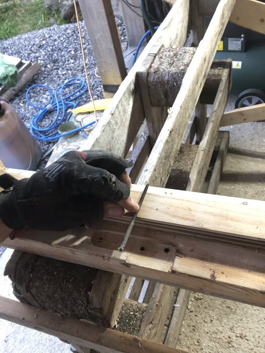 En person med en handske håller en lång skruv vid en del av en dörrkarm, omgivningen visar en verkstad med verktyg och virke.