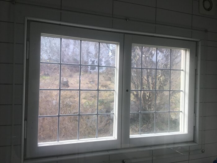 Ett dubbelfönster med frostat glas som släpper in ljus, omgiven av vitt kakel i ett badrum.