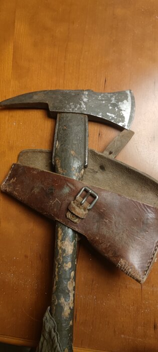 En sliten yxa med trähandtag och metallhuvud ligger på ett träbord, delvis täckt av ett gammalt läderfodral med spänne.