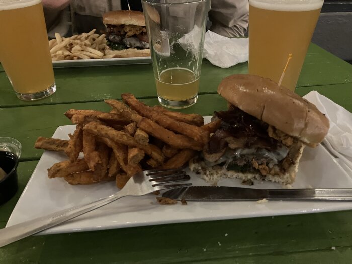 Hamburgare med bacon och ost på en tallrik, sötpotatispommes bredvid, glas med ljus öl i bakgrunden på ett grönt bord.