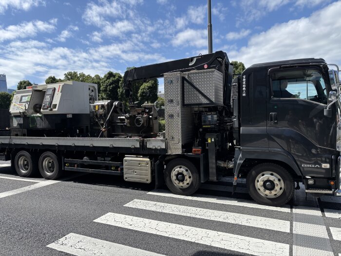 Svart lastbil med kran fraktar industriell maskin över övergångsställe i stadsmiljö, klar dag med blå himmel.
