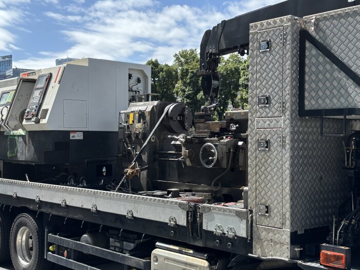 En CNC-maskin och annan utrustning på en lastbil under transport i centrala Tokyo, med vyer av stadens byggnader och träd i bakgrunden.