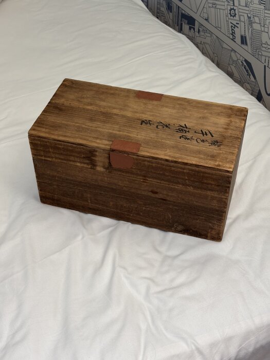 En träbox med mörk finish och handskriven text på locket, placerad på en vit säng.