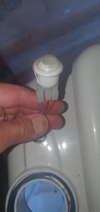 En hand håller i en vit plastkontakt med röda, blå och bruna kablar, bredvid en plastbehållare med ett runt hål.