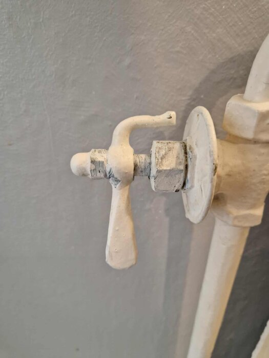 Gammal vitmålad slottsventil med invändig gänga, sitter monterad på ett rör mot en grå vägg.