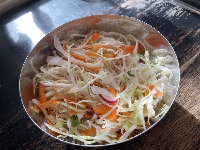 Metallskål med strimlad kålsallad, innehållande vitkål, morötter, rädisor och salladslök på ett mörkt underlag.