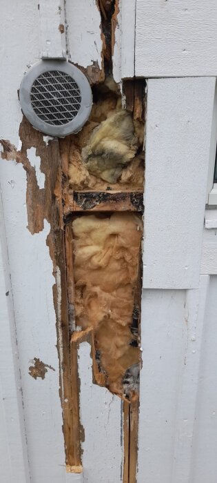 Exponerad väggisolering med skadat trä bakom trasig fasadbeklädnad, ventilationstrumma synlig upptill.