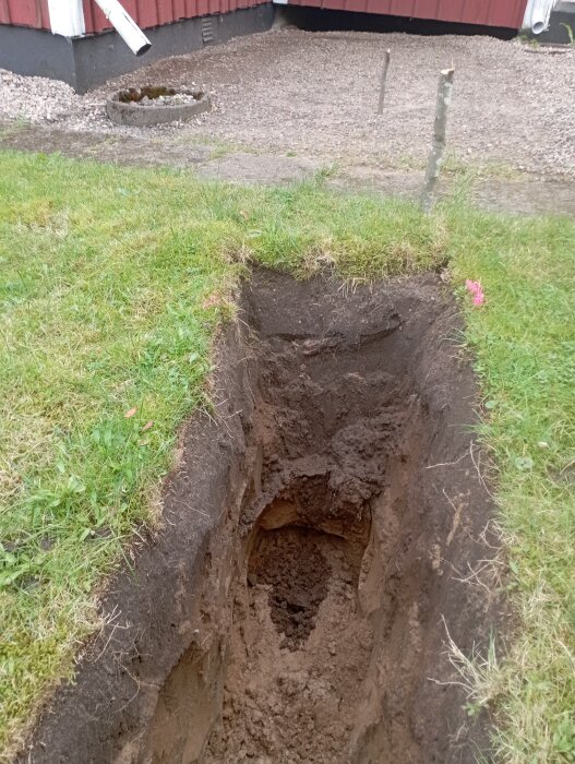 Grävd grop vid ett torp, visar jord och gräsmatta ovanpå. Synligt är husets hörn med röd fasad och en stenlagd gång.