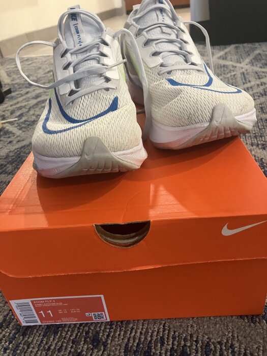 Ett par vita träningsskor med blå detaljer står ovanpå en orange skokartong på en grå matta.