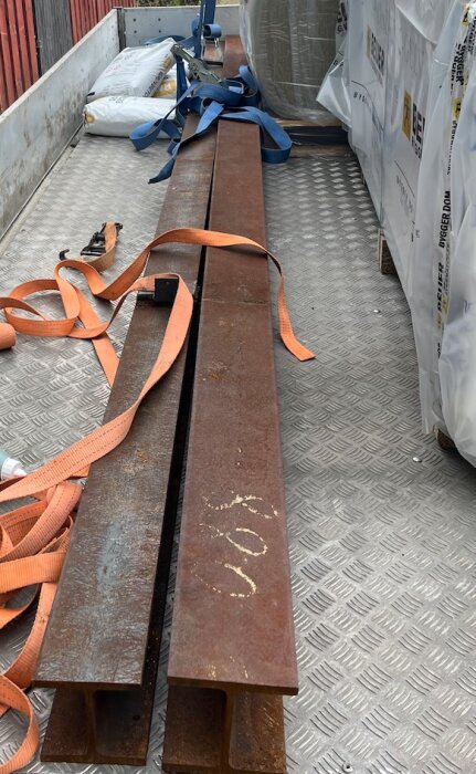 Stålbalkar ligger på ett lastbilsflak, säkrade med spännband för transport till ett byggprojekt i källaren.