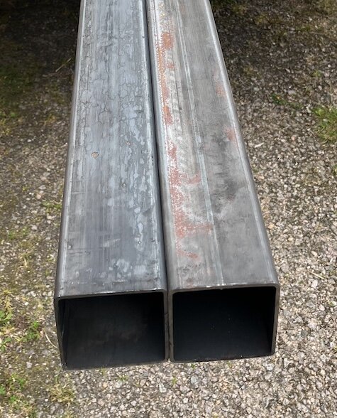 Två stålh-balkar ligger på grus, redo för installation som pelare i en källare.