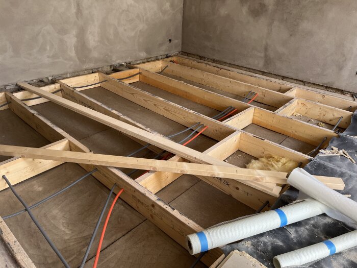 Golvbjälklag med öppna träbjälkar, kablar och byggmaterial synliga, förberedelse för ny isolering och golvläggning i ett renoveringsprojekt.
