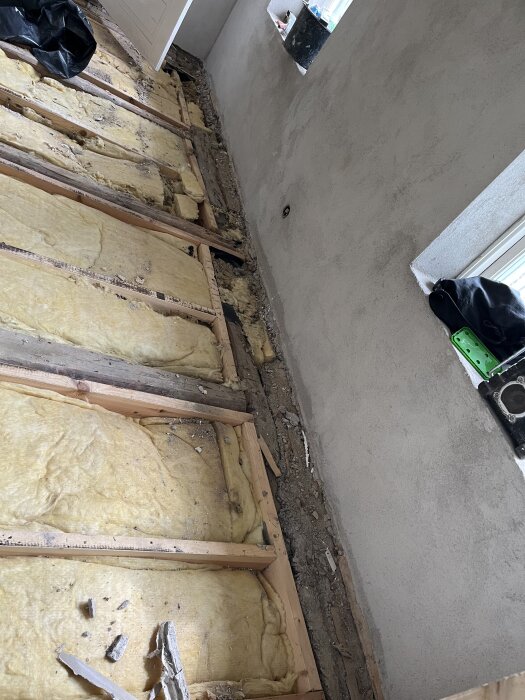 Öppnat golvbjälklag med isolering synlig, omgivna av väggar som nyligen blivit putsade. Byggmaterial på golvet.