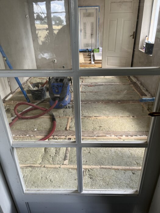 Rum under renovering, golvet öppnat för installation av ny isolering. Inga möbler, en dammsugare och verktyg syns i förrummet.