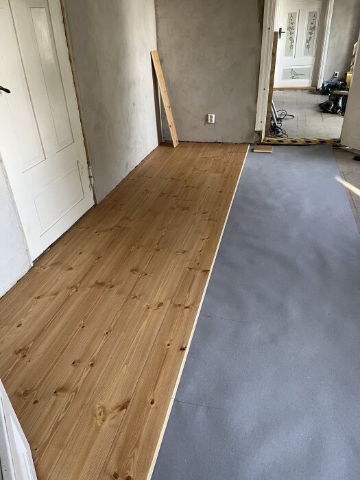 Nylagt trägolv i ett renoveringsprojekt, halvt lagd med underlagspapp synlig bredvid, dörröppning i bakgrunden och ofärdiga väggar.