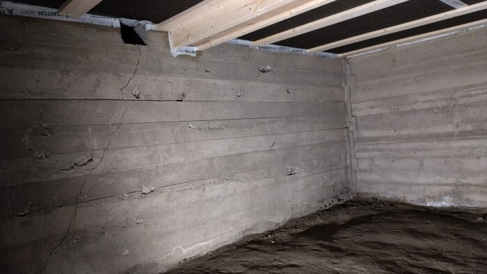 Rå betongvägg med sprickor och jordgolv i en oinredd källare. Trätakstolar syns längst upp i bilden.