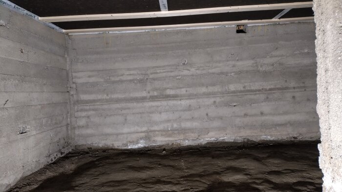 Betongvägg med jordgolv i källare, synligt utan plastskydd. Takhöjd cirka 160 cm. Används för lagring; funderingar på att gjuta betonggolv.