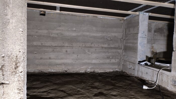 Betongväggar i en källare med jordgolv, synliga kablar och en liten hylla i hörnet. Ingen plast synlig.