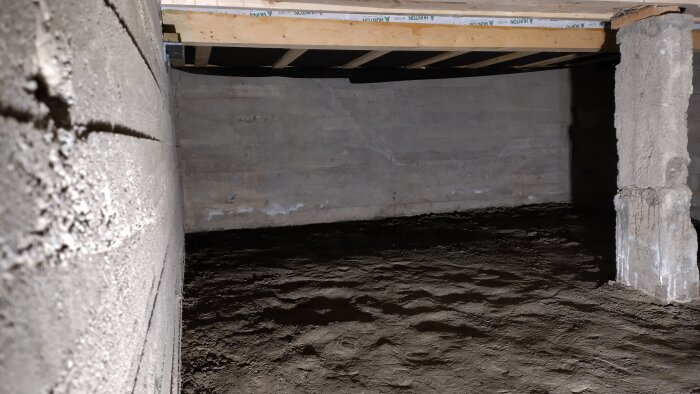 Bild av en källare med jordgolv, synliga betongväggar och -pelare. Träkonstruktioner syns i taket. Källaren används för lagring.