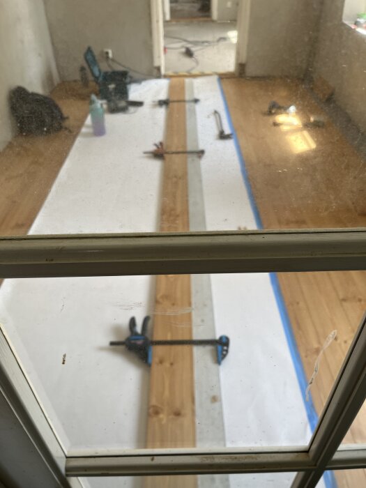 Vy genom dörr över pågående golvläggning med verktyg och pappskydd på plats i renoverat rum.