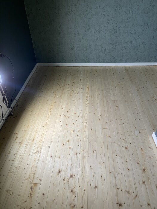 Ljust trägolv i rum mot blommig mörkgrön tapet, efter slipning och behandling.
