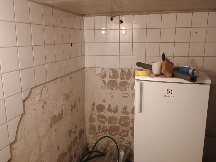 Gammal källarvägg med kakel och avflagande puts, på golvet står ett kylskåp och diverse byggmaterial ovanpå.