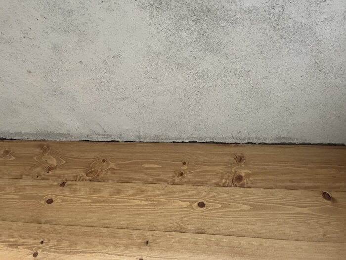 Trägolv med tydliga kvistmärken möter en vitmålad väggyta; synlig liten spalt mellan golvbrädor och vägg.
