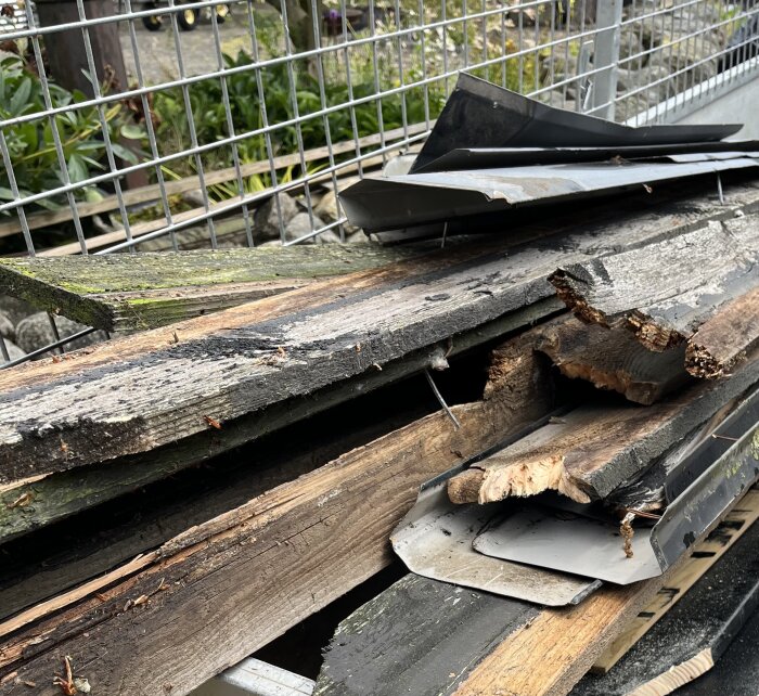 Rivna träplankor och plåtstycken från takkupor ligger i en hög bredvid ett staket.