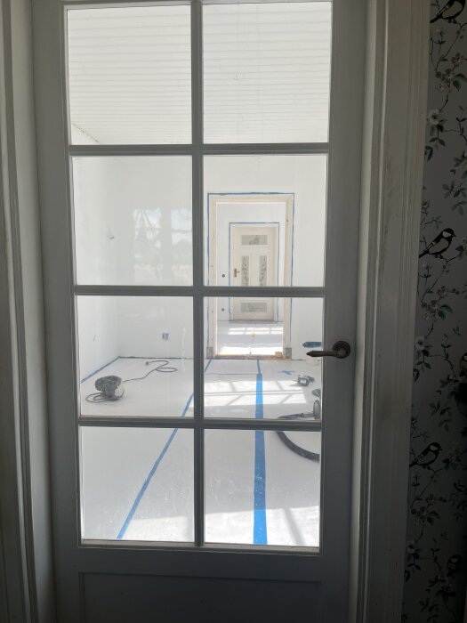 Vy genom glasdörr mot ett rum med nyligen vitmålade väggar och tak. Golvet skyddat med papp och blå tejpremsor syns, verktyg ligger på golvet.